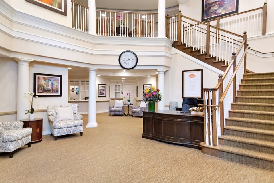 A spacious and elegant senior living facility lobby with beige carpet and cream-colored walls. There is a dark wooden reception desk with a black office chair behind it and a vase of colorful flowers on top. To the left, there are two patterned armchairs with a small round wooden table between them, holding a white orchid plant. The lobby features white columns and a staircase with wooden handrails and carpeted steps leading to an upper level with a white railing. Several framed paintings and a large round clock are mounted on the walls.