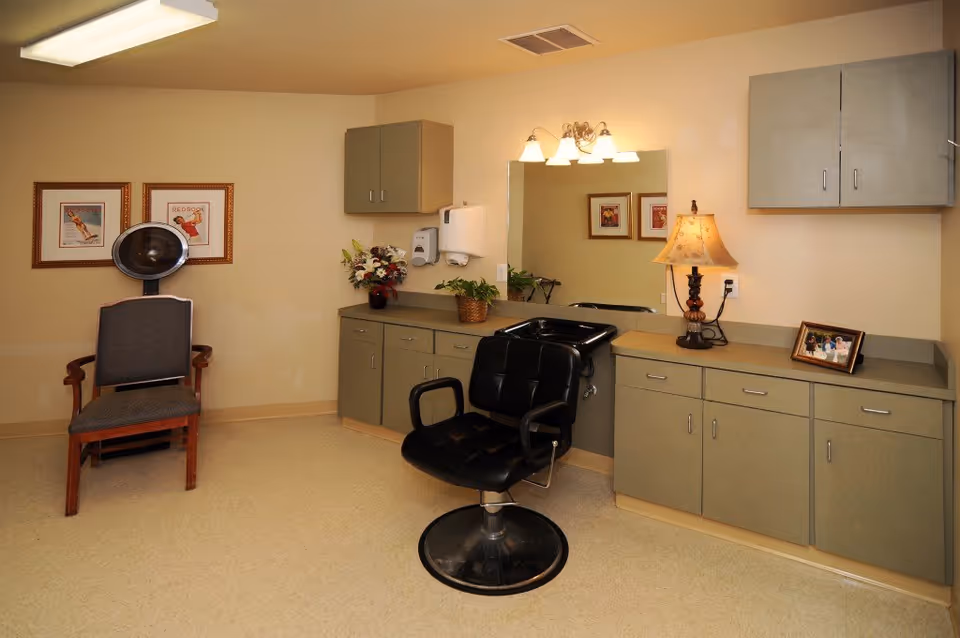 Salon-style interior with a black barber chair by a sink, cabinets, a hair dryer chair, framed pictures and a lamp.