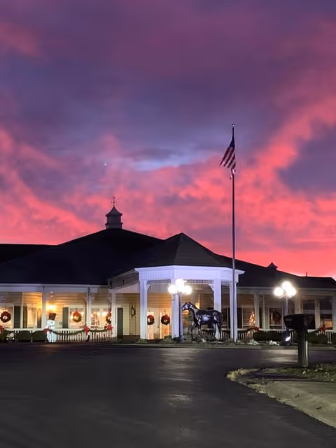 Front entrance of a single-story assisted living building at dusk with a flagpole, a horse statue, wreaths in the windows, and a vivid pink-purple sky.