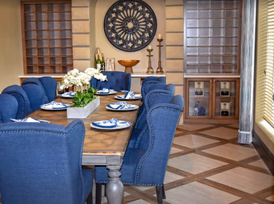 A dining room with a wooden table set for six, featuring blue upholstered chairs with nailhead trim. The table is decorated with a rectangular planter of white flowers and blue and white napkins on plates. The room has a patterned tile floor, two windows with wooden blinds, a decorative round wall piece, and a shelf with candles and bottles.