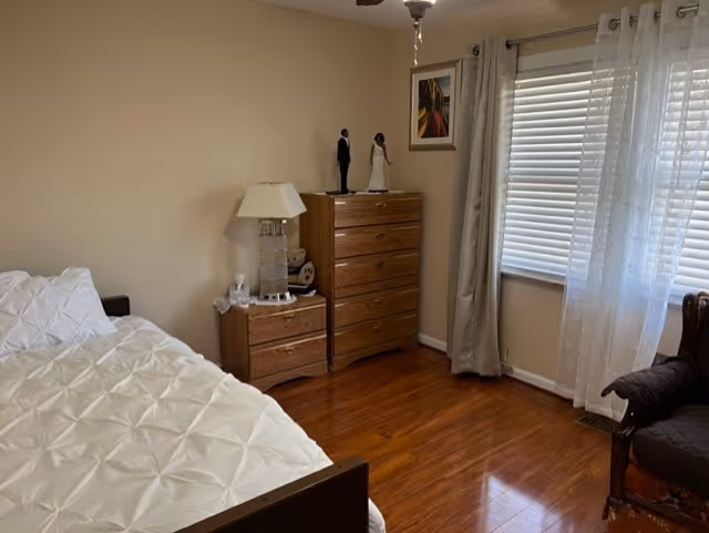 Bedroom with a bed covered in a white comforter, wooden nightstand and dresser, a window with blinds and curtains, and hardwood floors.
