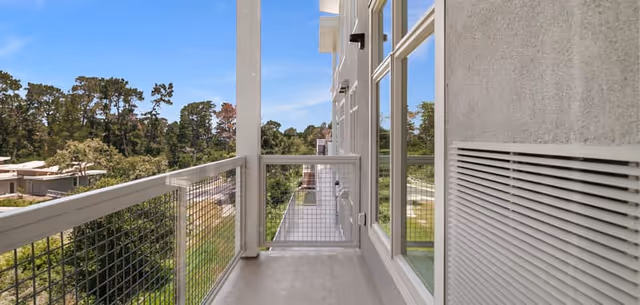 A narrow balcony with a metal railing and mesh panels attached to a building. The balcony overlooks a green area with trees and other buildings in the distance under a clear blue sky.