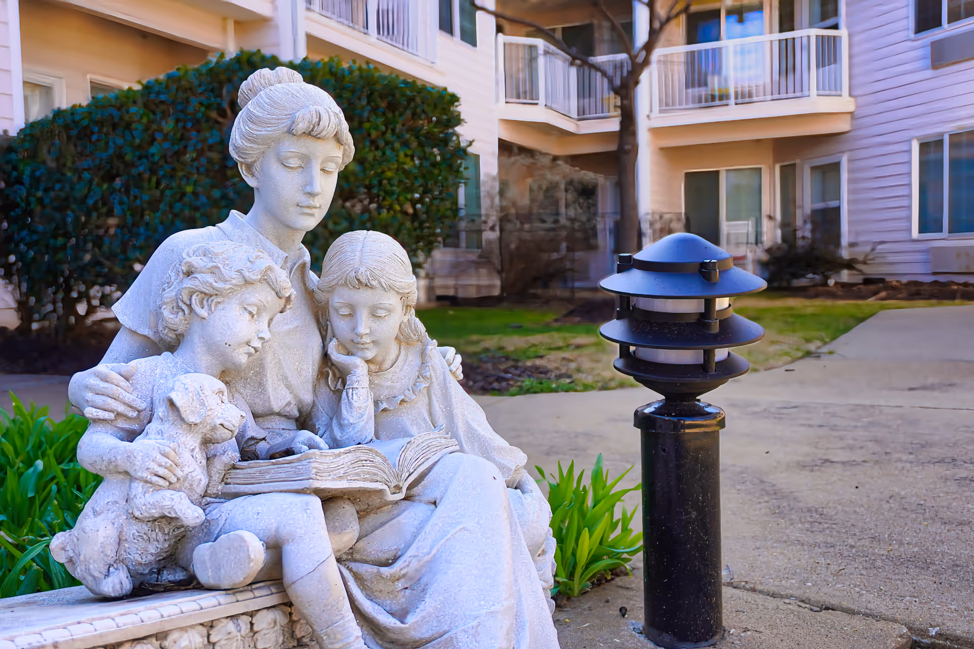 A stone statue of a woman reading a book to two children, one holding a small dog, situated outdoors near a pathway with a modern black lamp post and residential building in the background.