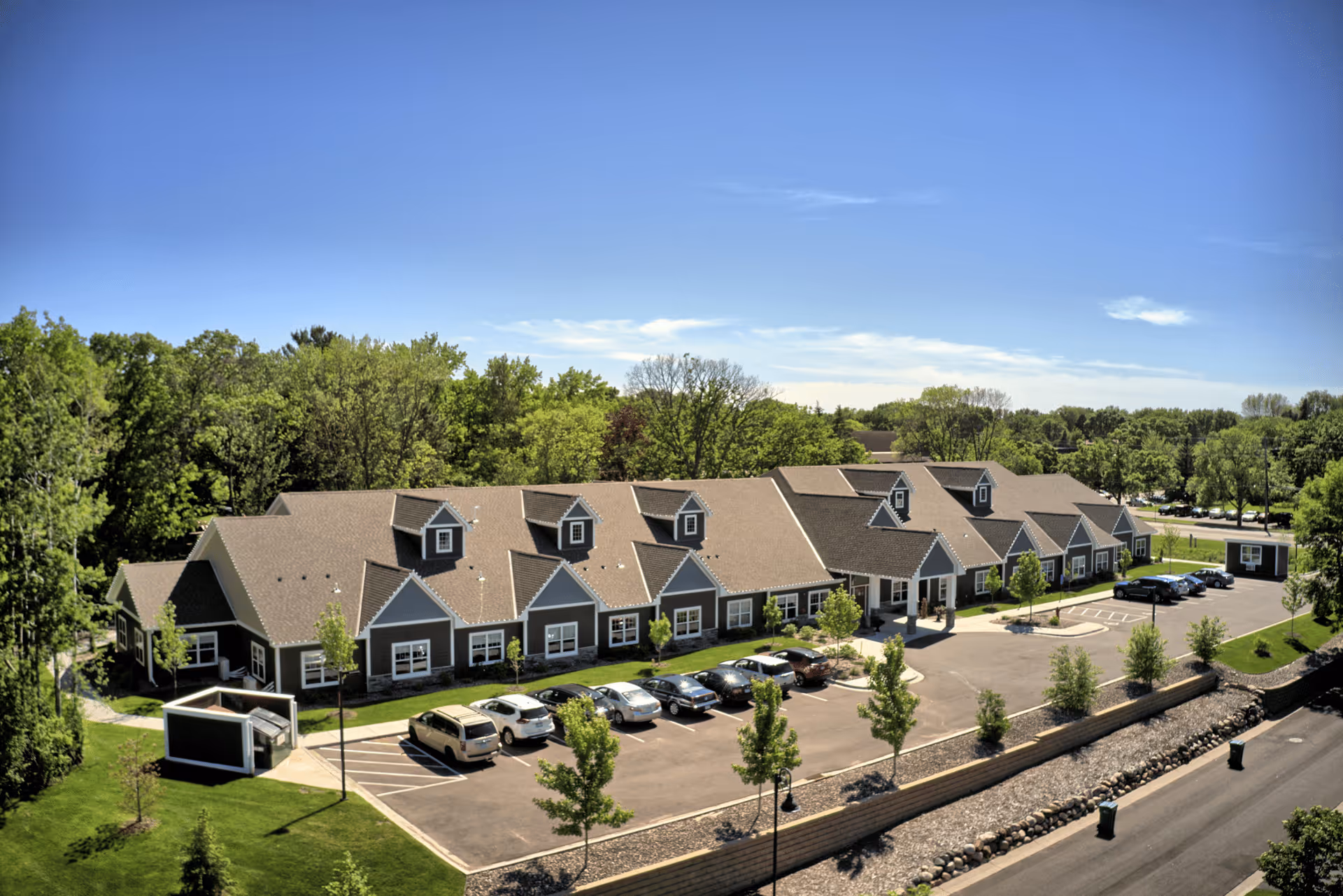 Aerial view of a single-story senior living facility building with a brown roof and multiple dormer windows. The building is surrounded by green trees and landscaping, with a parking lot in front containing several parked cars. The sky is clear and blue.