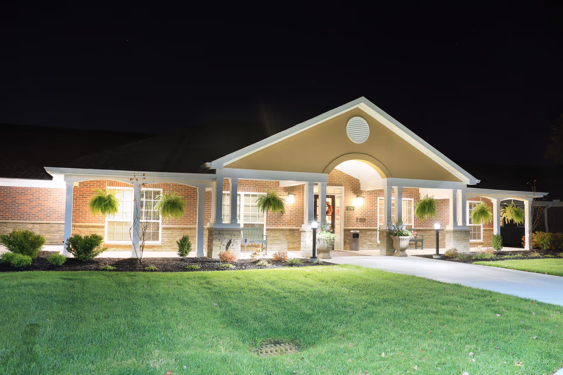 Night view of the front exterior of a senior living facility building with a well-lit entrance, hanging plants, shrubs, and a green lawn.