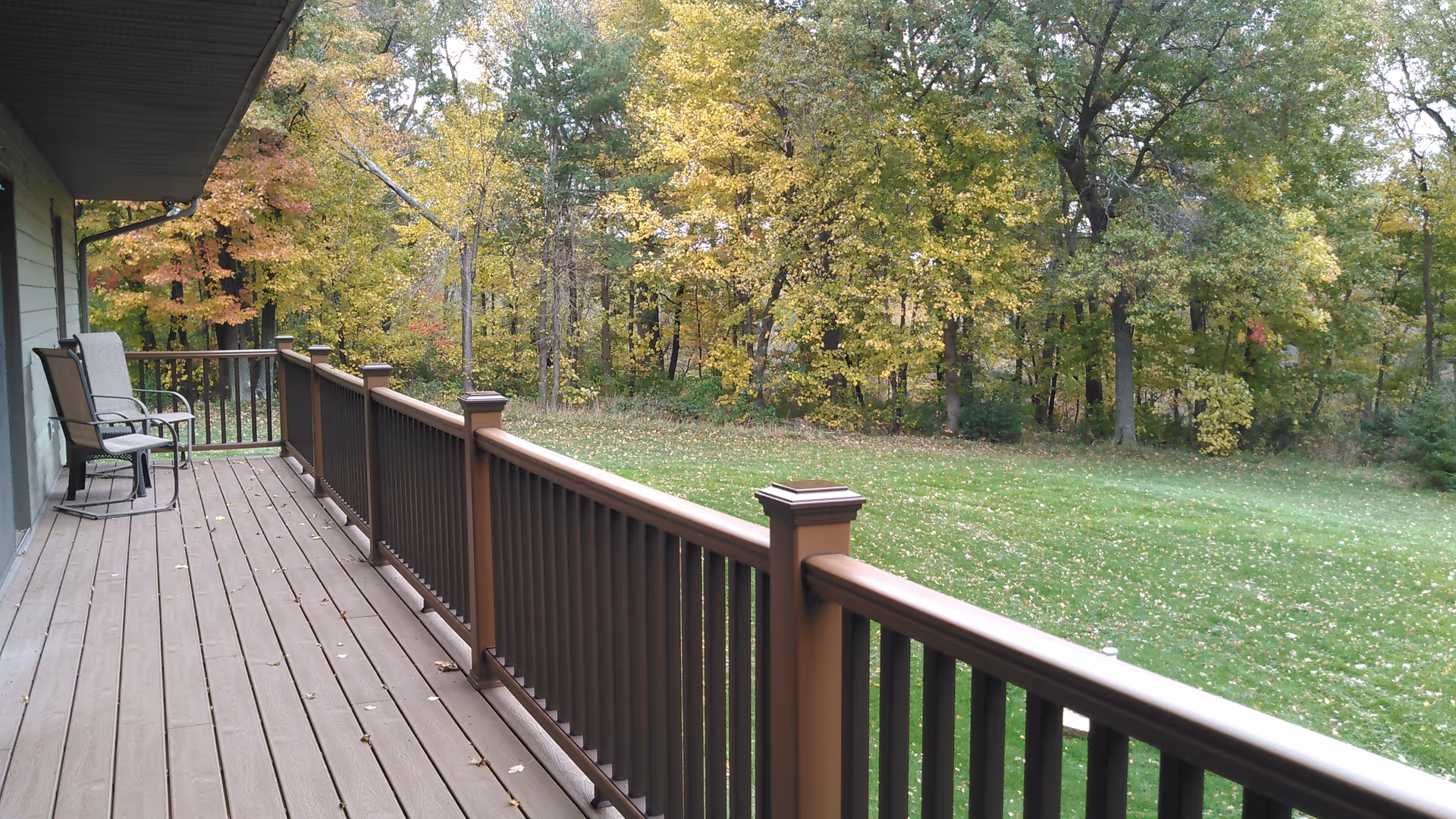 A wooden deck attached to a building with two chairs and a small table. The deck overlooks a grassy yard and a forested area with trees showing autumn foliage.