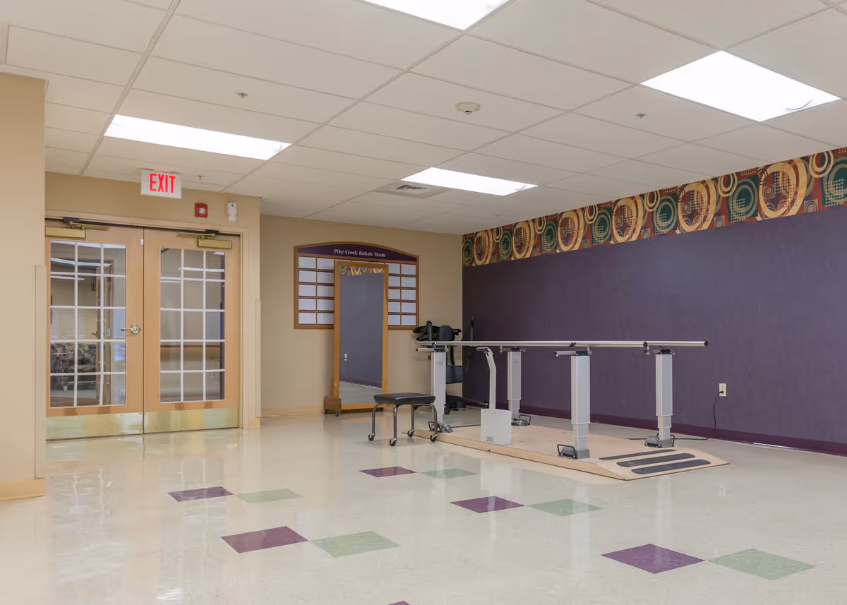 Spacious rehabilitation room with parallel walking bars, a rolling stool, purple accent wall and double doors under an EXIT sign.