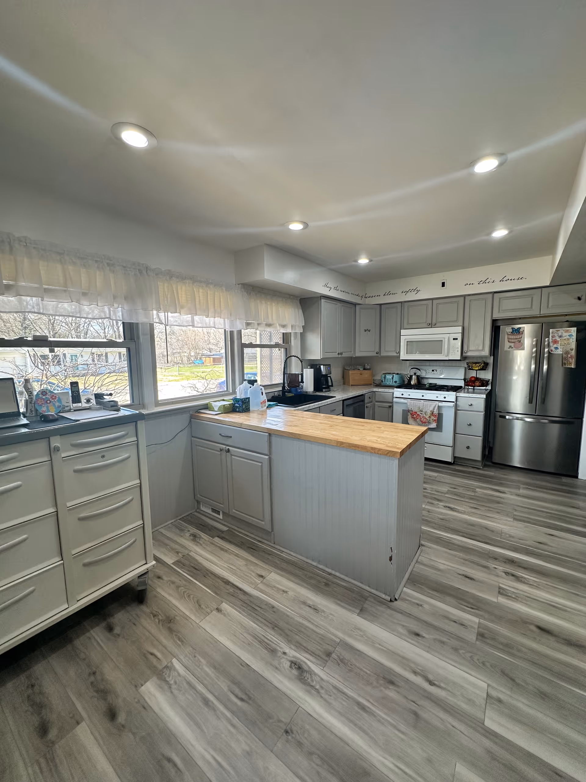 A bright and modern kitchen with light gray cabinets, a wooden countertop island, stainless steel refrigerator, white microwave, and stove. The kitchen has large windows with sheer white curtains letting in natural light, and the floor is covered with gray wood-like planks. Recessed ceiling lights illuminate the space.