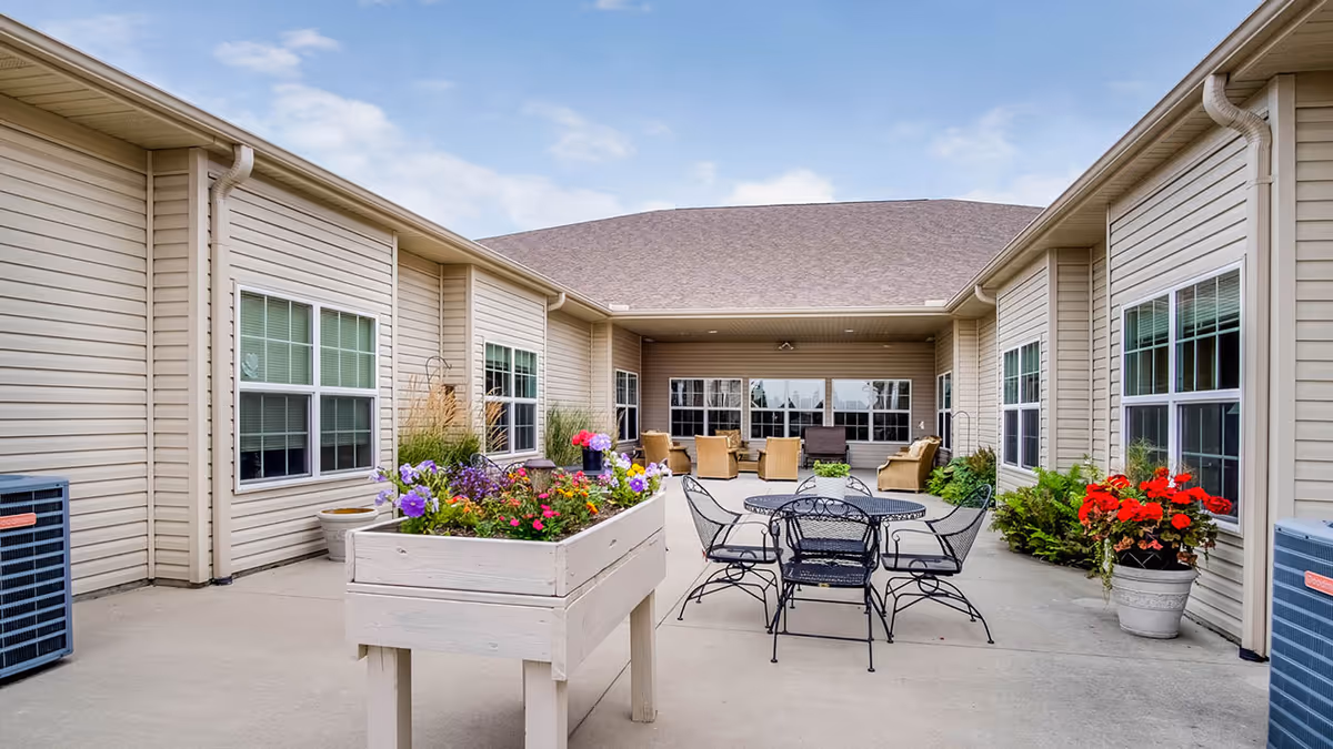 Sunny courtyard patio at the assisted living facility with metal tables and chairs, lounge seating, and colorful flower planters between beige vinyl-sided building wings.