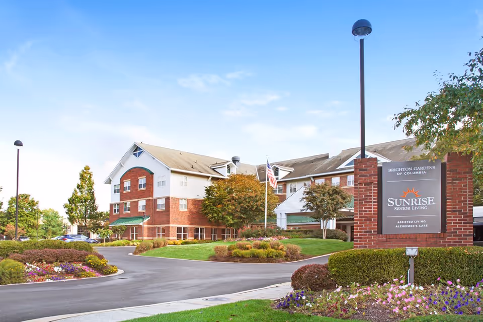 Exterior view of Brighton Gardens of Columbia, a senior living facility with a brick and white building, surrounded by landscaped gardens, trees, and a driveway. A sign in front reads 'Brighton Gardens of Columbia, Sunrise Senior Living, Assisted Living Alzheimer's Care.'