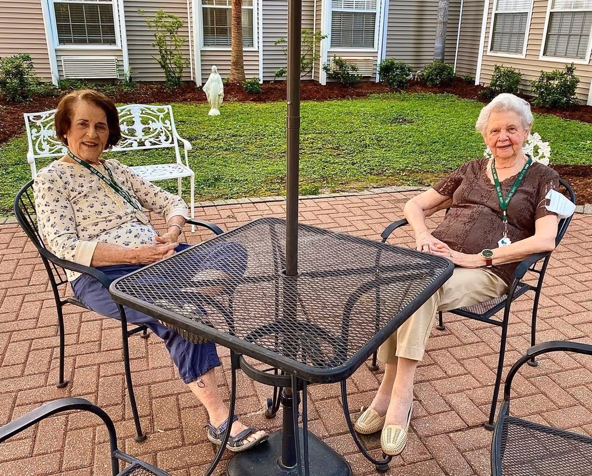 Two elderly women sitting outdoors at a black metal patio table with chairs on a brick-paved area. Behind them is a green lawn with bushes and a white statue near the building with beige siding and white window shutters.
