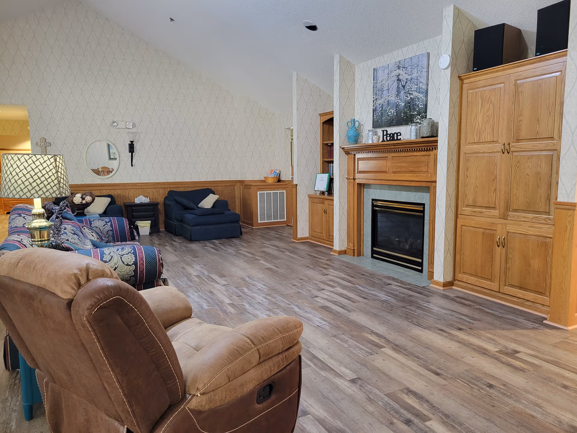 A cozy living room in an assisted living facility featuring a brown recliner chair, a patterned sofa, a dark blue armchair, a wooden fireplace with decorative items and a painting above it, built-in wooden cabinets, a floor lamp, and wood-patterned flooring.