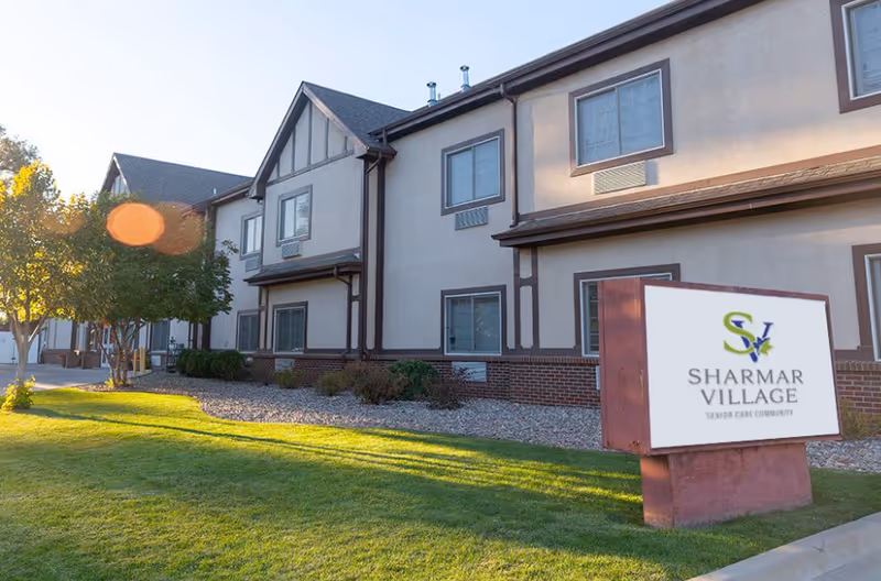 Exterior view of a two-story senior care community building with beige walls, brown trim, and multiple windows. In front of the building is a green lawn, some trees, and a sign that reads 'Sharmar Village Senior Care Community'.