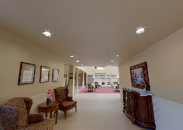 A hallway in an assisted living facility with beige walls and ceiling lights. On the left side, there are two upholstered armchairs with a small wooden table between them, decorated with a flower arrangement. Three framed pictures hang on the wall above the chairs. On the right side, there is a wooden cabinet with a framed picture above it. The hallway opens up to a larger room with carpeted flooring and additional seating visible in the background.