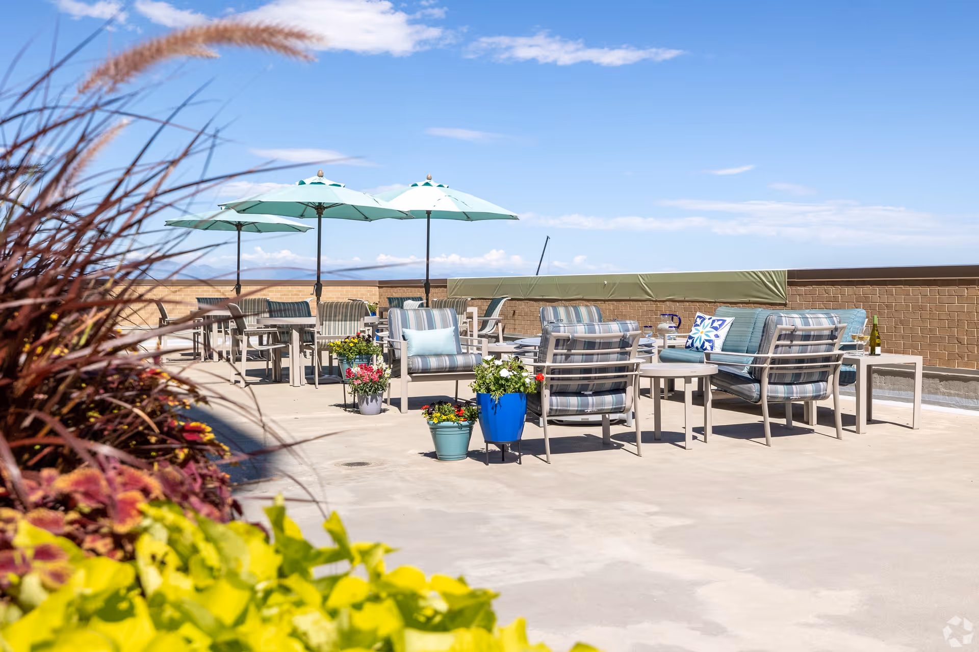 Outdoor patio area with several seating arrangements including cushioned chairs and sofas with striped upholstery, small tables, and umbrellas providing shade. There are colorful potted plants in the foreground and a clear blue sky with some clouds in the background.