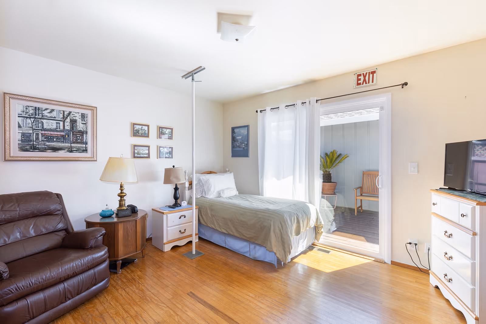 A bright and cozy bedroom with a single bed covered in light green bedding, a brown leather recliner chair, a wooden side table with a lamp, a white nightstand with another lamp, and a white dresser with a TV on top. The room has wooden flooring, several framed pictures on the walls, and a sliding glass door with white curtains leading to an outdoor patio area with a chair and a potted plant. An exit sign is visible above the door.