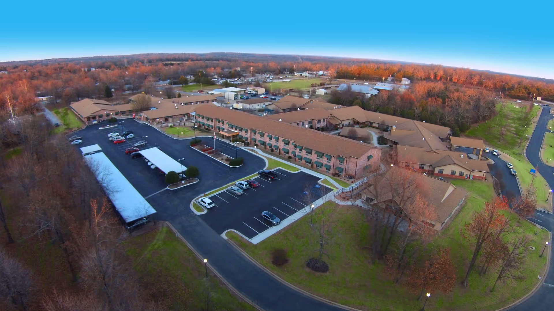 Aerial view of Lake Forest Senior Living at Mountain Home showing a large senior living facility with multiple connected buildings, parking lots with several cars, surrounding trees with autumn foliage, and nearby roads.