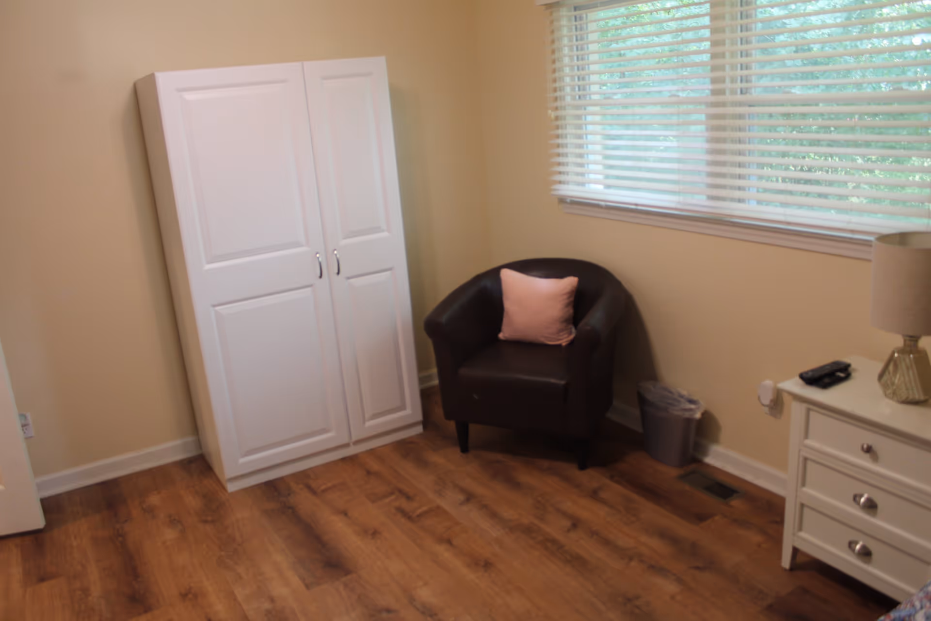Small room with a white wardrobe, a brown armchair with a pink pillow, a white nightstand and lamp, and wood-look flooring under a window with blinds.