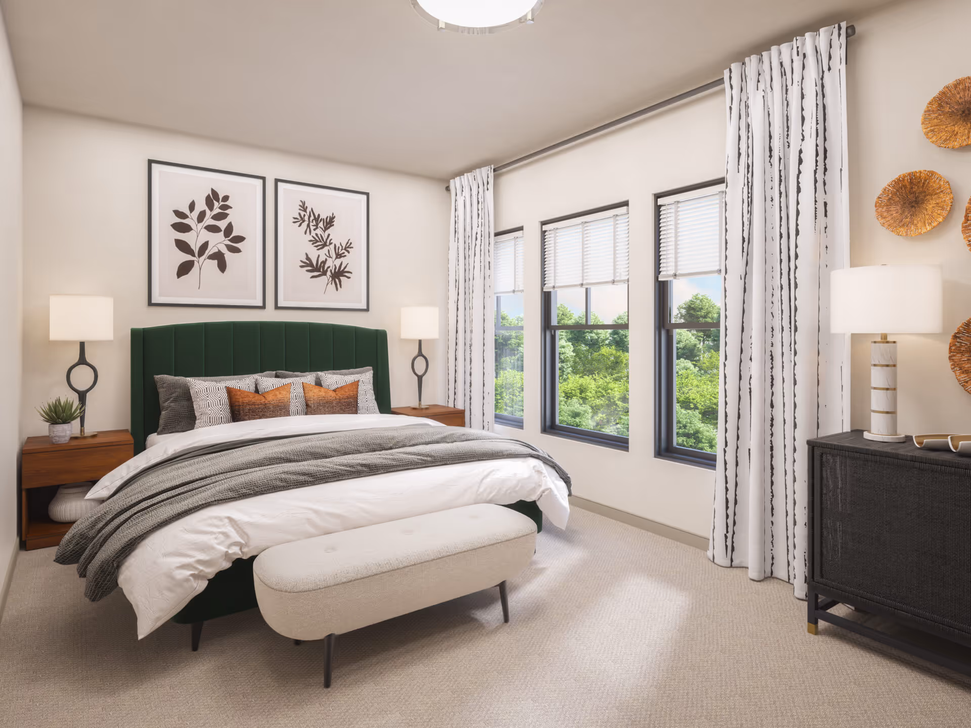 A bright and cozy bedroom with a green upholstered bed, white bedding, and multiple pillows. There are two framed botanical prints on the wall above the bed, two wooden nightstands with modern lamps, a small plant on one nightstand, and a cushioned bench at the foot of the bed. Three large windows with white blinds and patterned curtains let in natural light, and a black dresser with a lamp and decorative items is on the right side of the room.