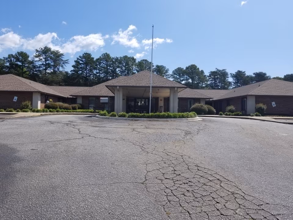 Front exterior view of a single-story brick building with a covered entrance and a flagpole in front. The building is surrounded by trees and has a cracked asphalt driveway leading up to the entrance.