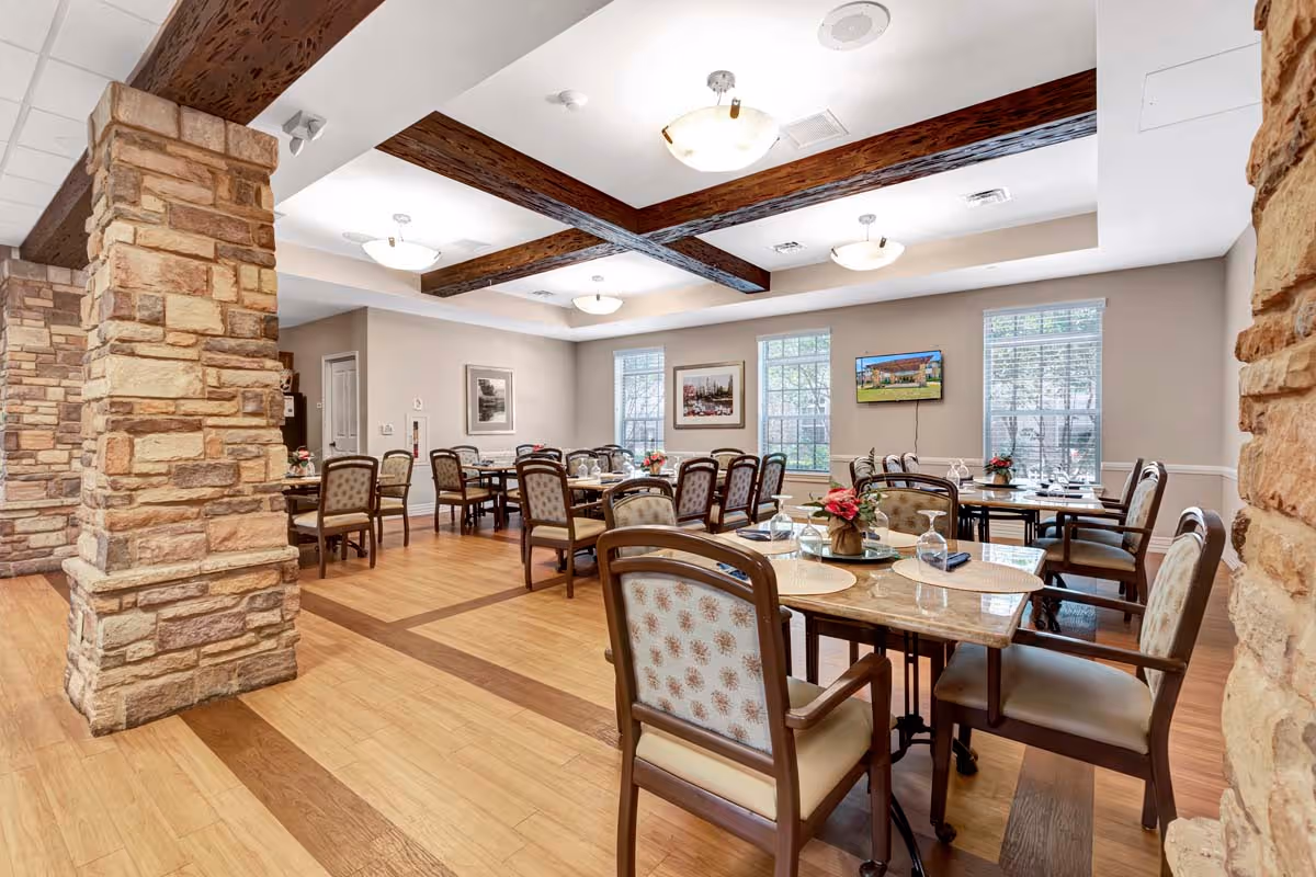 A spacious dining room with multiple tables and chairs arranged neatly. The room features wooden beams on the ceiling, stone pillars, large windows letting in natural light, and framed artwork on the walls. Tables are set with placemats, glassware, and small floral centerpieces.