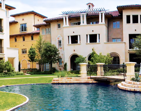 Exterior view of a multi-story residential building with beige and light brown walls, surrounded by green trees and shrubs, with a small bridge over a pond in the foreground.