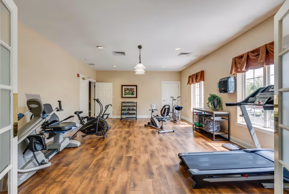 A bright exercise room with wooden flooring featuring various fitness equipment including a treadmill, stationary bikes, and a rack of dumbbells. The room has beige walls, two windows with brown valances, and a small table with plants and exercise accessories.