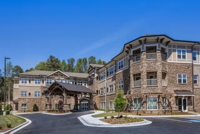 Exterior view of a multi-story senior living facility building with stone and beige siding, multiple windows, balconies, and a covered entrance under a clear blue sky.