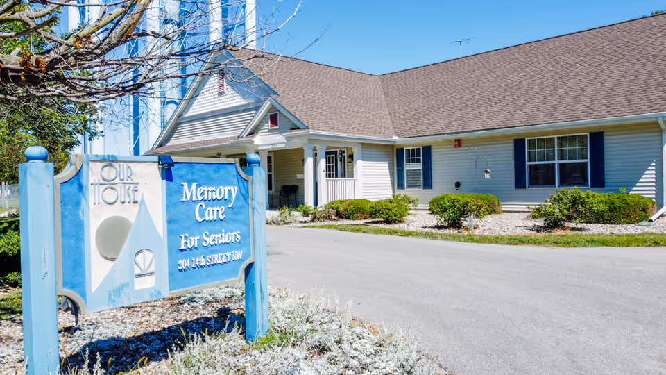 Exterior view of a single-story senior living facility building with beige siding and a brown roof under a clear blue sky. In front of the building is a blue and white sign that reads 'Our House Memory Care For Seniors 204 14th Street NW'. There are some bushes and landscaping around the building and sign.