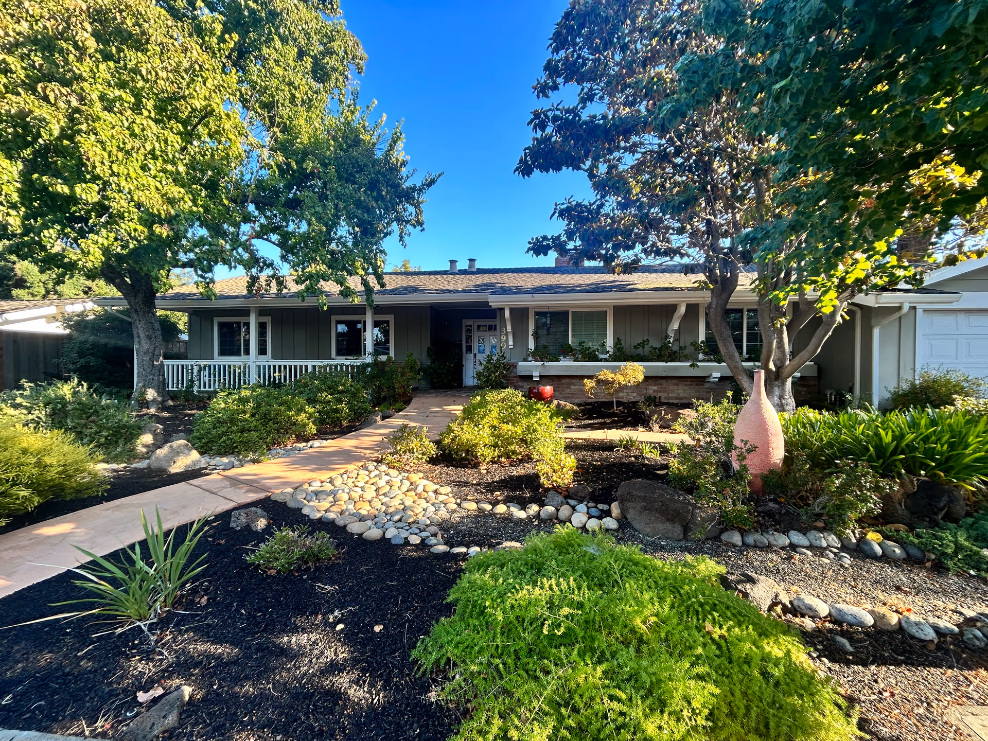 Single-story ranch-style home with a front porch, landscaped yard, walkway, and trees under a clear blue sky.