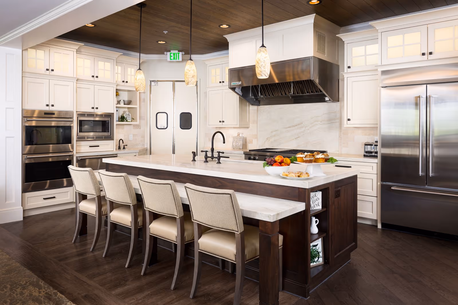 A modern kitchen with white cabinetry, stainless steel appliances including a double oven and large refrigerator, a central island with a dark wood base and white marble countertop, four beige cushioned chairs, pendant lights hanging from a wooden ceiling, and a bowl of fruit and pastries on the island.