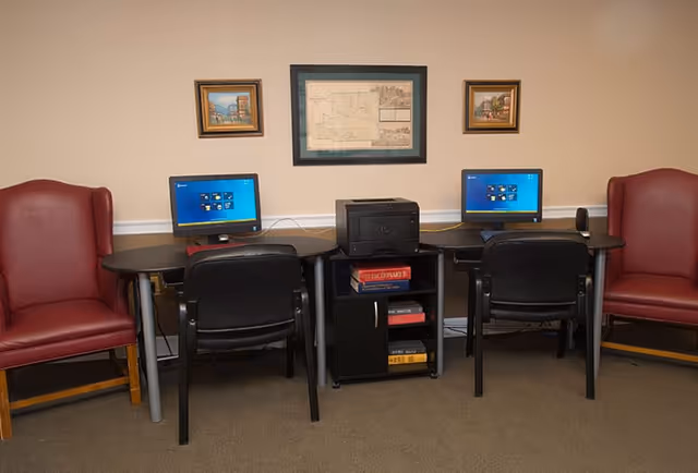 A small computer workstation area with two desktop computers on black desks, two black chairs, a black printer on a small shelf unit with books, and two red armchairs on either side. Three framed pictures hang on the beige wall above the desks.