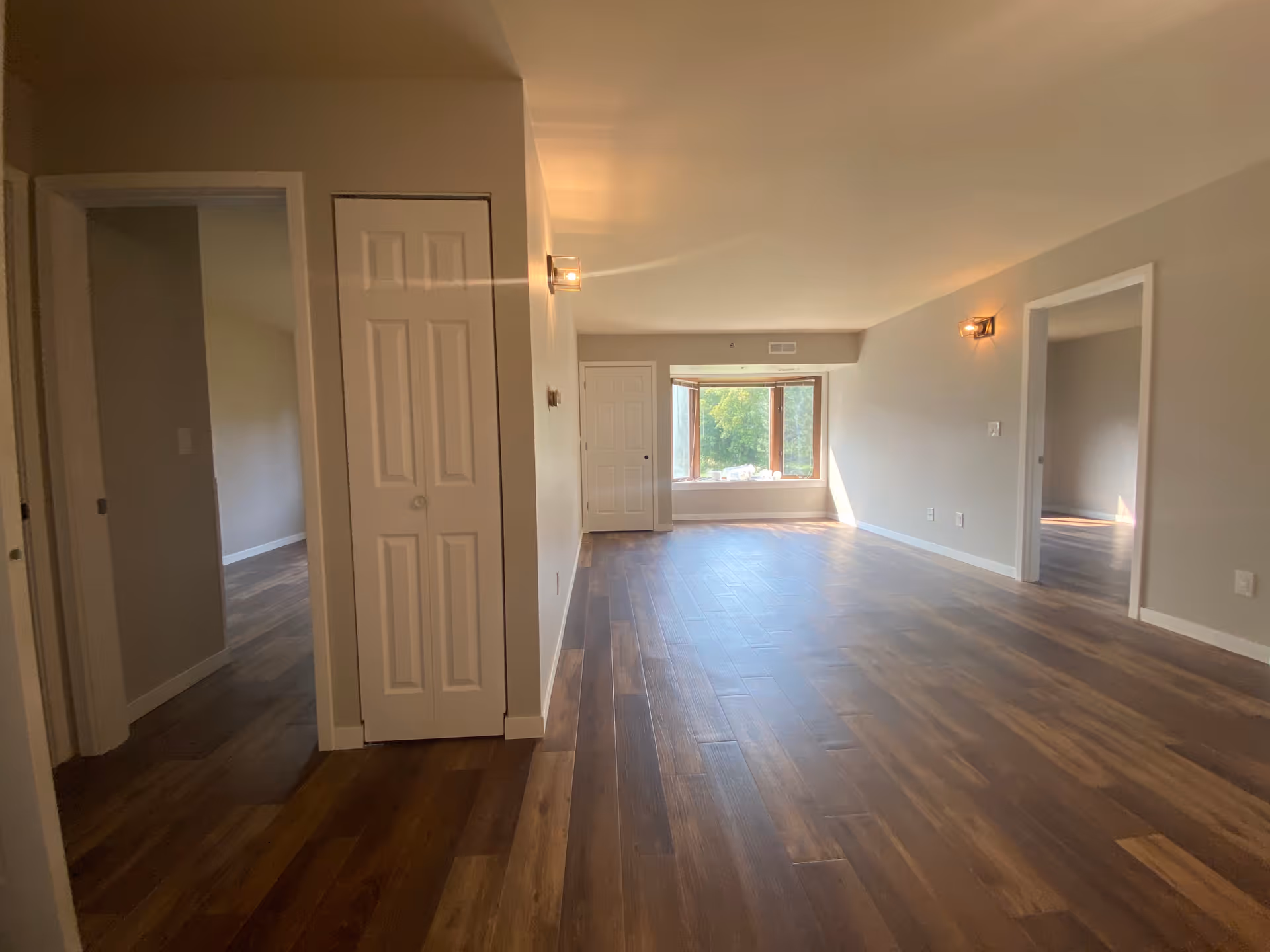 Empty interior room with wooden flooring, beige walls, two doorways leading to other rooms, a closet with white double doors, wall-mounted light fixtures, and a large window at the far end letting in natural light.