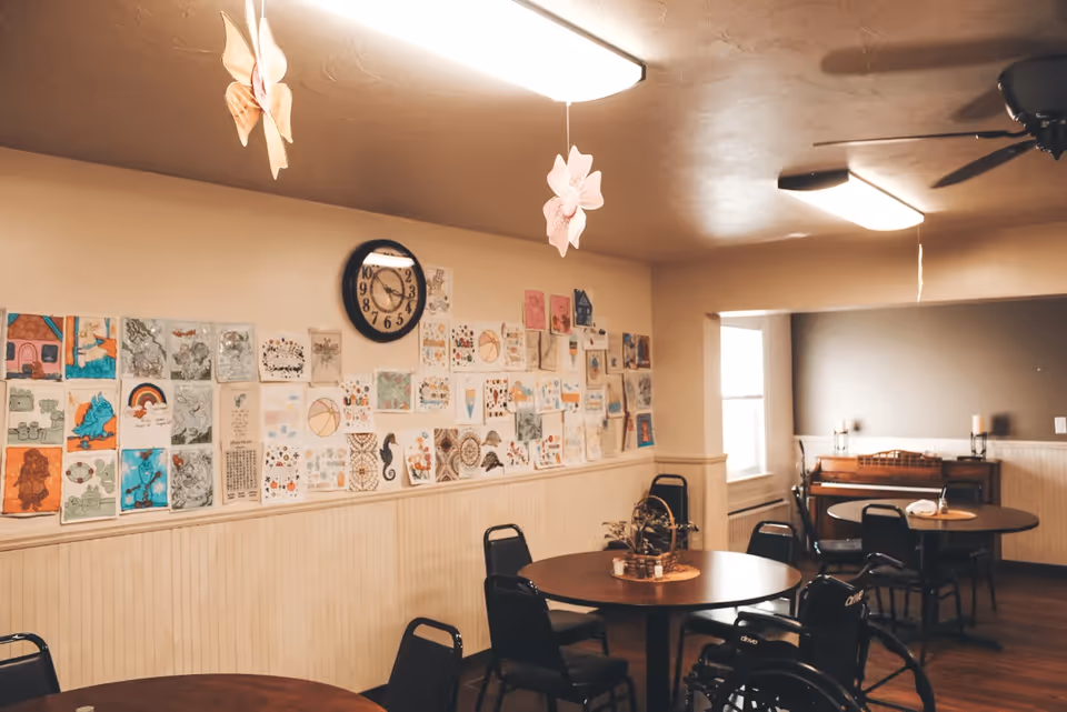 A cozy common room with round tables and chairs, a wheelchair, and a wall decorated with various colorful drawings and artwork. A clock is mounted on the wall above the artwork. The room has warm lighting with ceiling lights and hanging flower decorations.