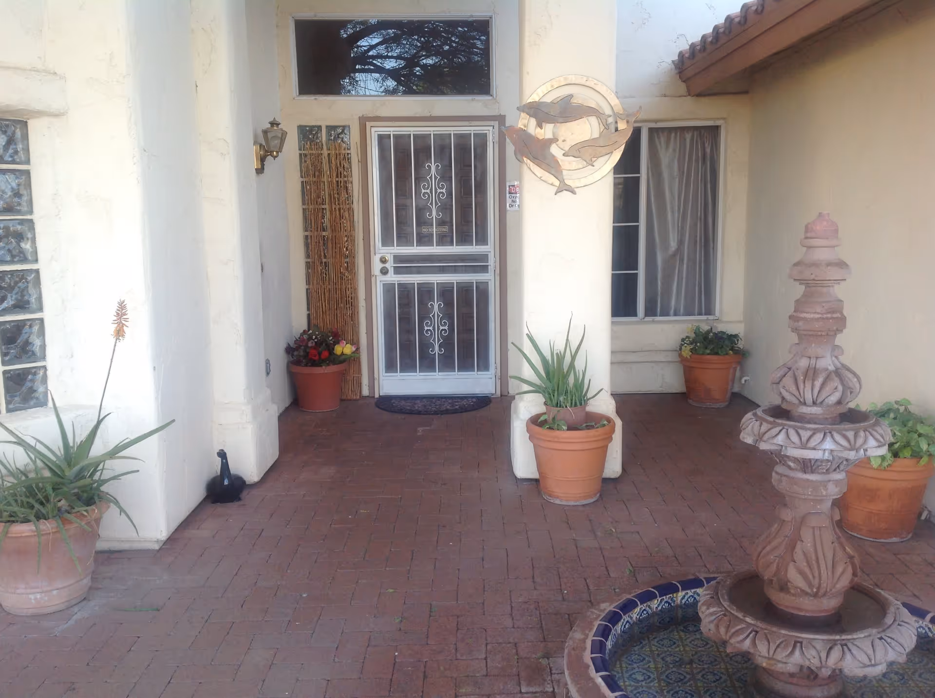 Front entrance of a building with a white security door, potted plants, a decorative wall hanging with dolphins, and a multi-tiered stone fountain on a brick patio.