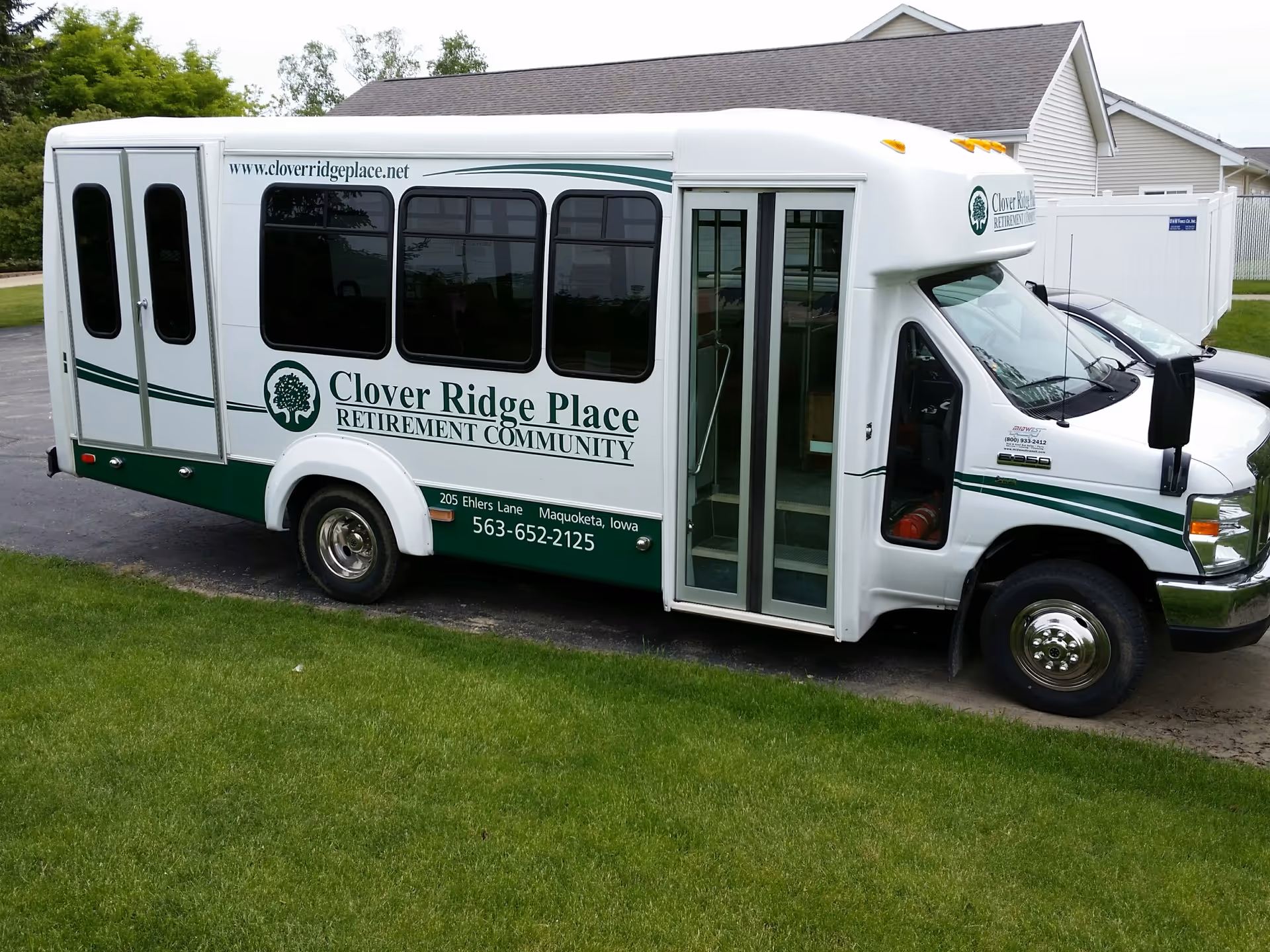 A green-and-white Clover Ridge Place retirement community shuttle bus parked on a driveway near houses and lawn.