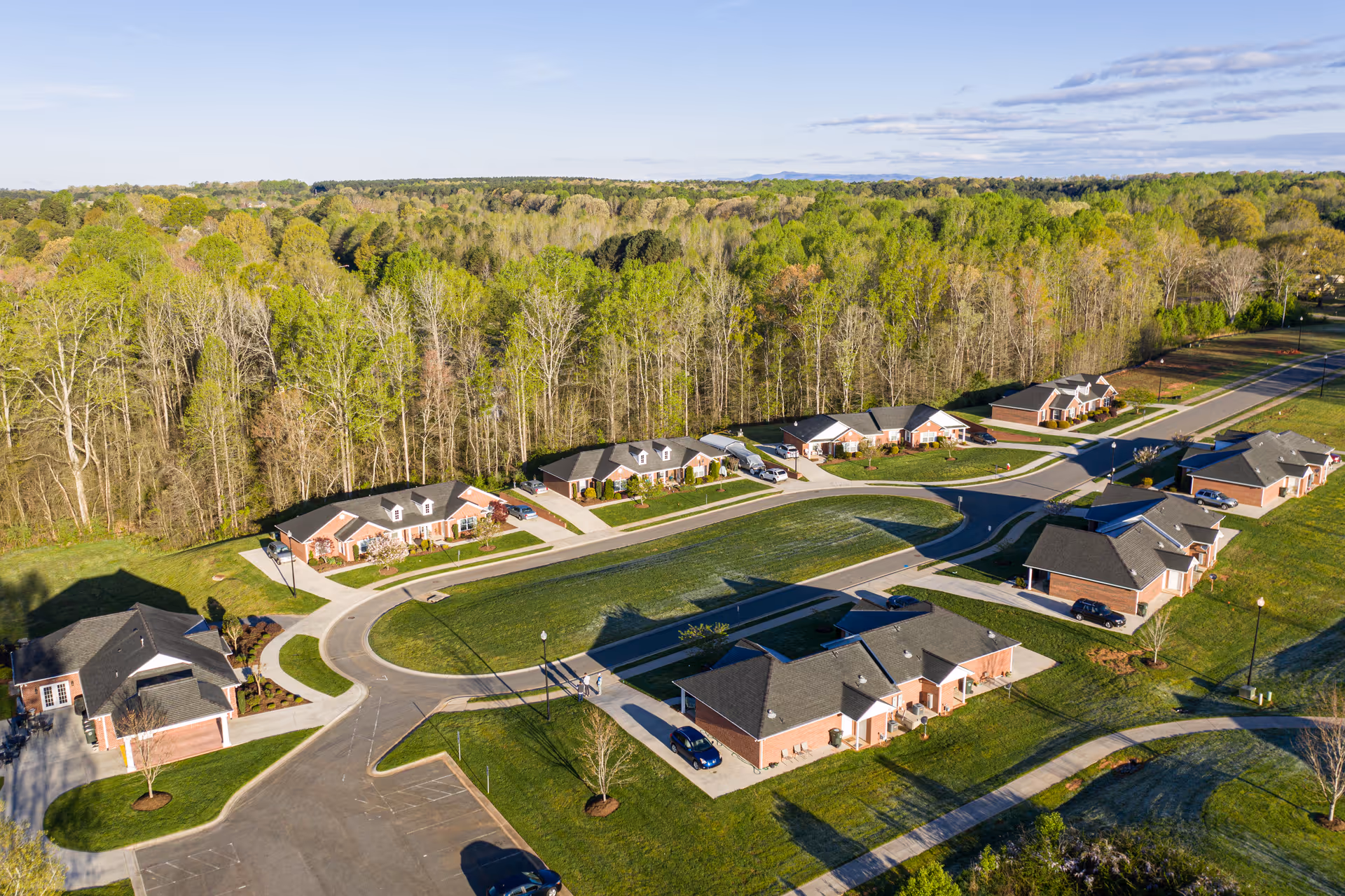 Aerial view of a retirement community with multiple single-story brick cottages arranged around curved roads and green lawns, bordered by a dense forest of trees under a clear sky.