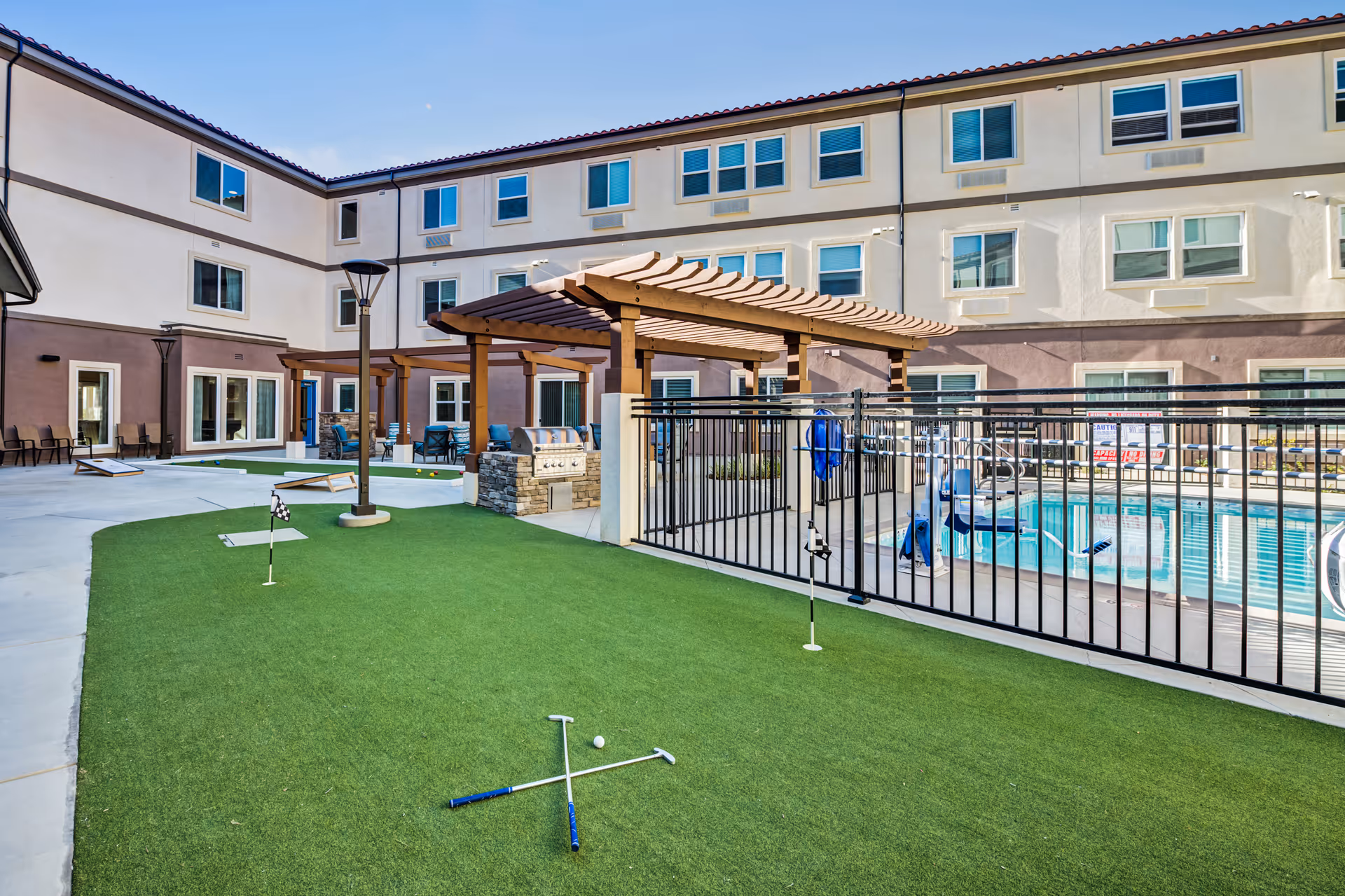 Outdoor courtyard area of a senior living facility with artificial putting green, two golf putters and a golf ball on the grass, a fenced swimming pool with a pool lift, pergolas with seating and a built-in barbecue grill, surrounded by a three-story building with multiple windows.