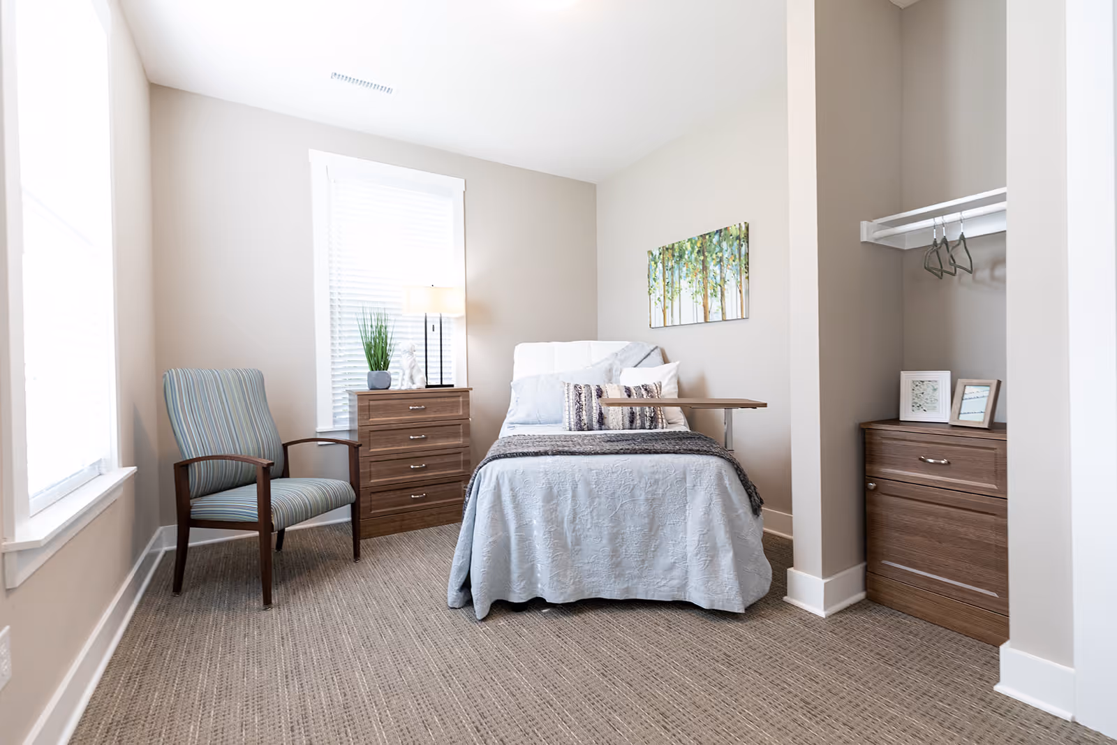 A bright and clean bedroom featuring a single bed with light blue bedding and multiple pillows. Next to the bed is a wooden dresser with a lamp and a plant on top. A striped armchair is positioned near a window with white blinds. On the right side, there is an open closet space with hangers and a small wooden cabinet with framed pictures. A colorful painting of trees hangs on the wall above the bed.