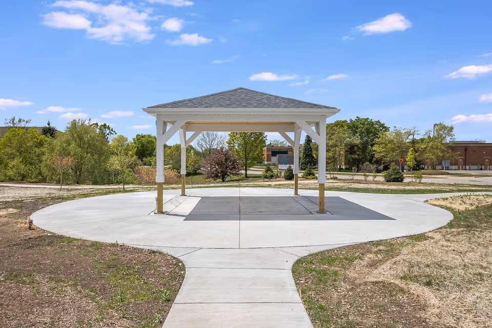 A small open pavilion with a gray shingled roof and white wooden supports stands on a circular concrete pad surrounded by a landscaped area with grass and trees under a blue sky with scattered clouds.