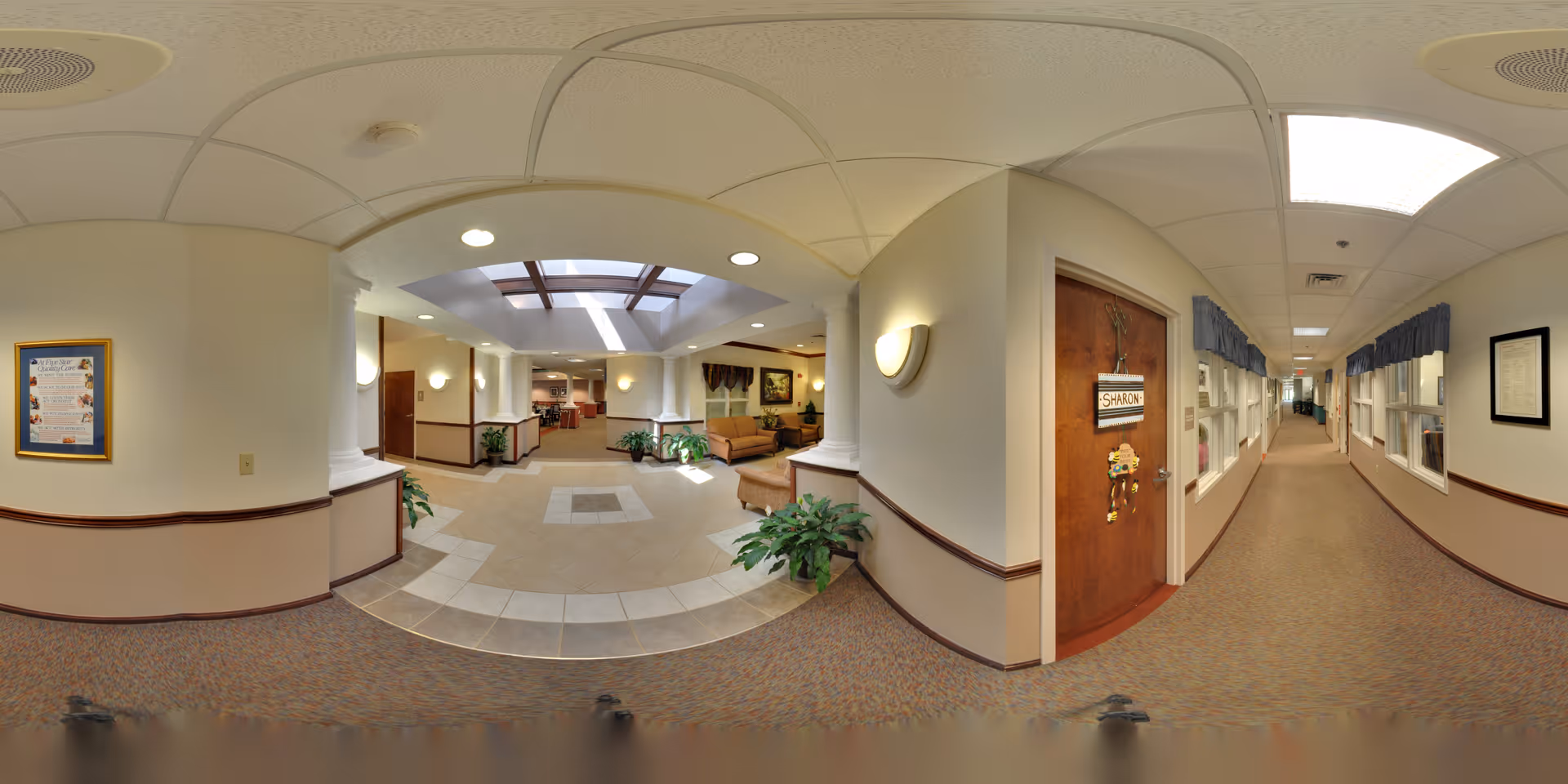 A wide panoramic view of a senior living facility interior hallway with a seating area under a skylight. The hallway has carpeted floors, beige walls with wooden trim, and several framed pictures. There is a wooden door labeled 'Sharon' with decorations, and windows with blue valances line the corridor. Potted plants are placed near the seating area and columns.