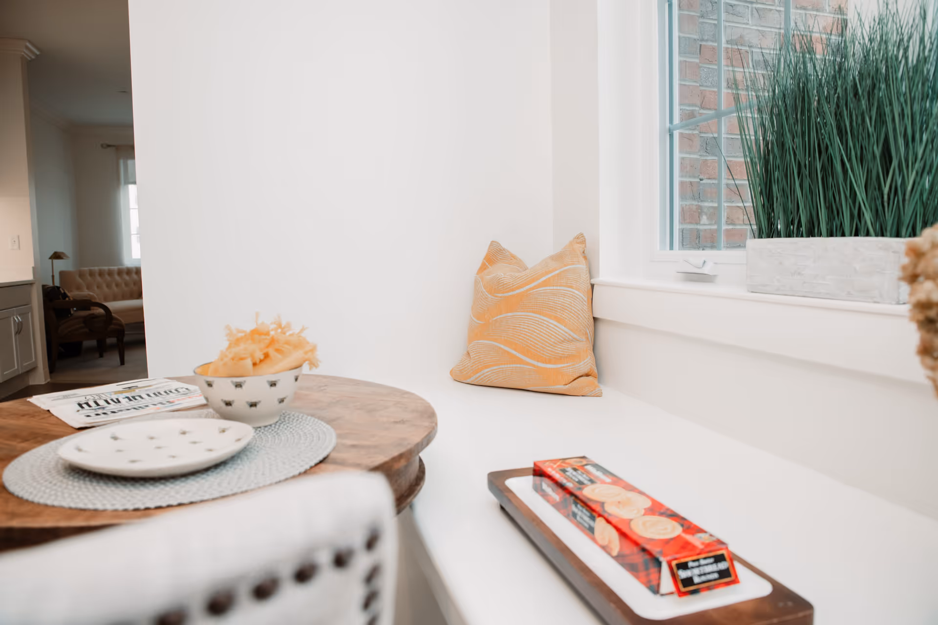 A cozy corner dining nook with a wooden round table set with a placemat, a bowl with decorative items, and a plate. There is a cushioned bench with an orange patterned pillow next to a window with a potted plant on the windowsill. In the background, a beige tufted sofa and a wooden chair are visible in an adjacent room.