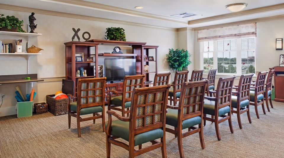 A well-lit room with rows of wooden chairs facing a large wooden entertainment center with a flat-screen TV. The room has beige walls, carpeted floor, and a large window with blinds letting in natural light. There are decorative plants, shelves with books and ornaments, and exercise equipment in the corner.