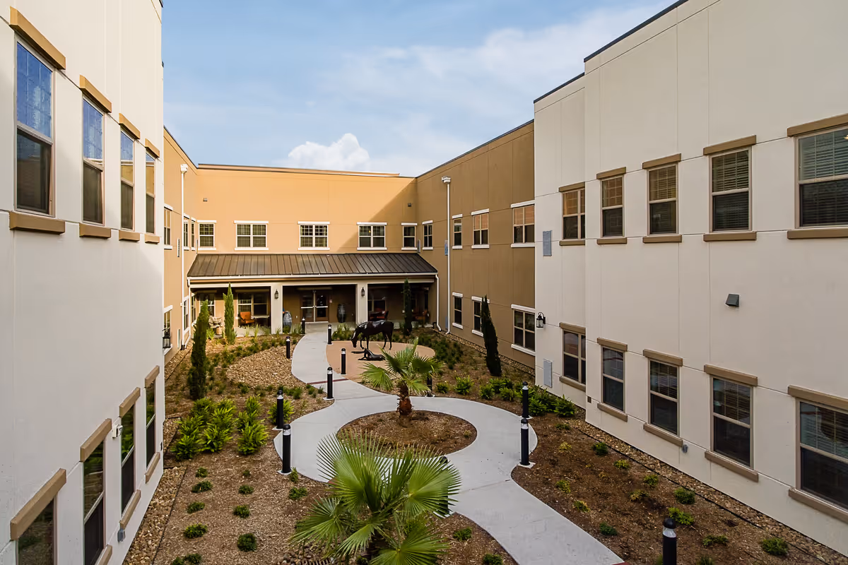 Outdoor courtyard area of a senior living facility with a winding concrete pathway, landscaped plants, small trees, and a horse statue in the center. The courtyard is surrounded by a two-story building with multiple windows under a partly cloudy sky.