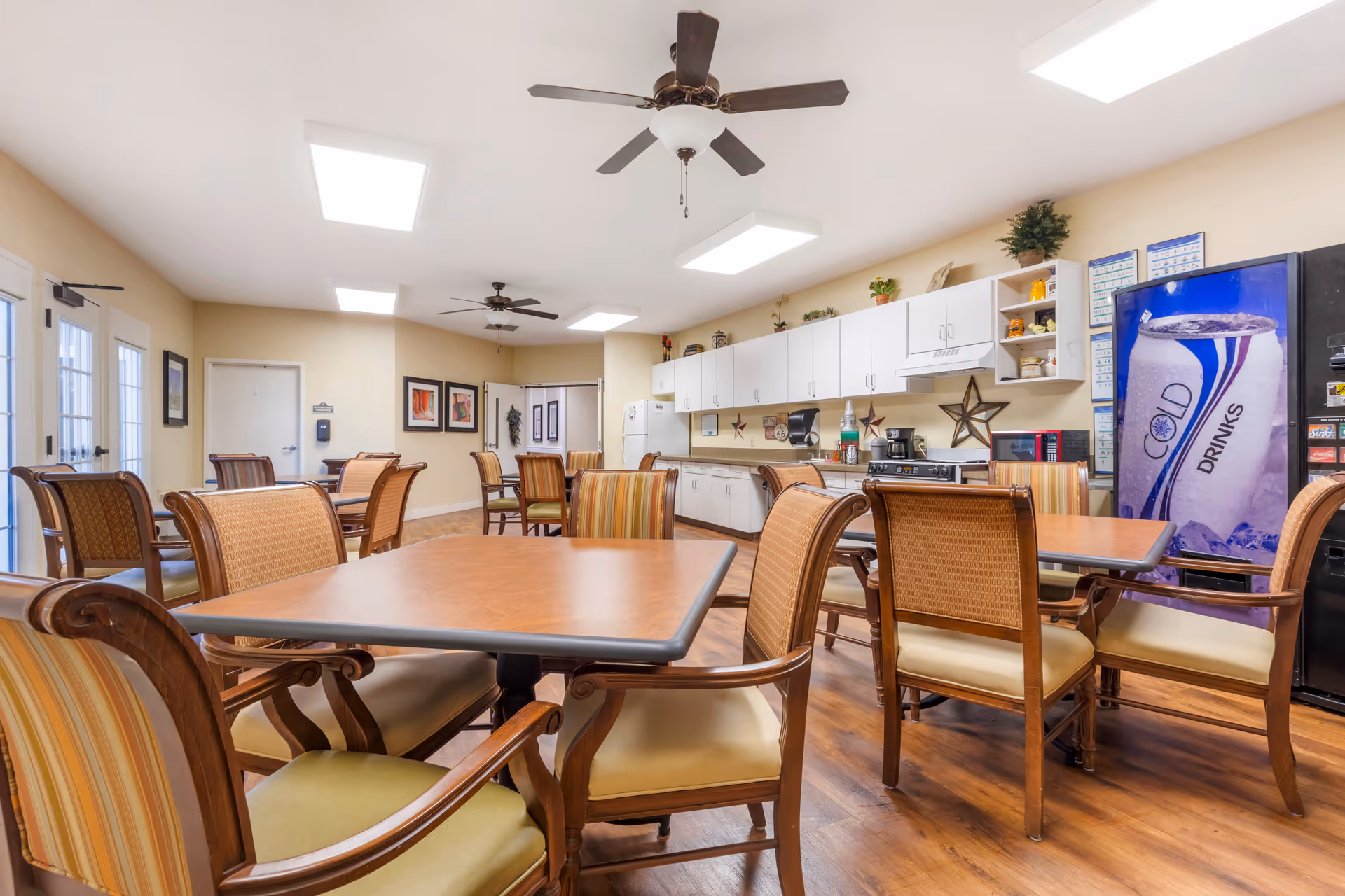 Community dining area with wooden tables and chairs, a kitchenette with white cabinets, ceiling fans, and a vending machine.