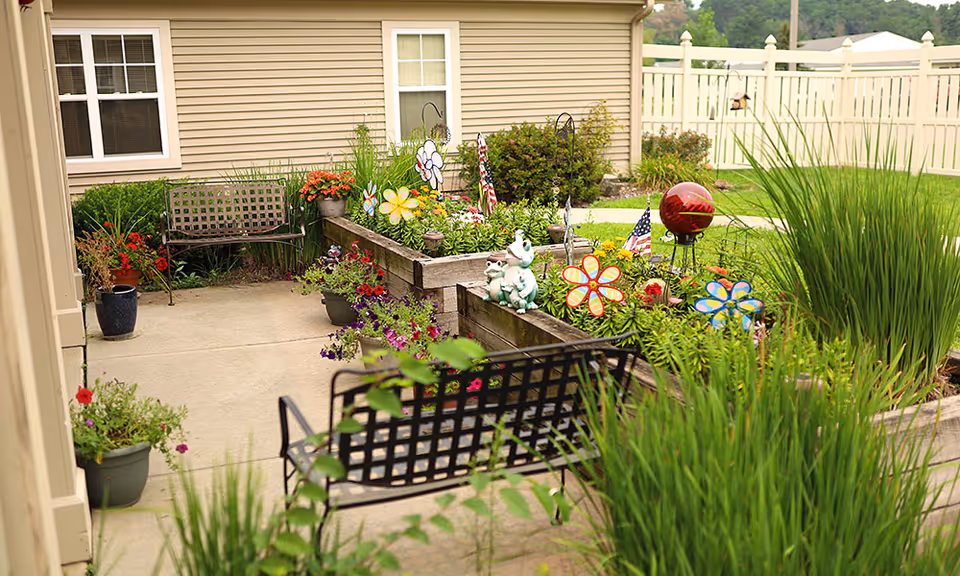 Outdoor garden area with raised flower beds filled with colorful flowers and decorative items including flower-shaped ornaments and small American flags. There are two black metal benches on a concrete patio next to a beige building with windows and a white fence in the background.