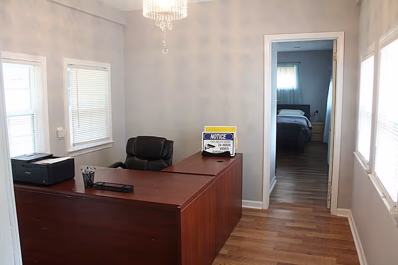 An office area with a wooden desk and a black office chair. On the desk, there is a printer, a pen holder with pens, and a sign that reads 'NOTICE THIS AREA IS UNDER 24 HOUR VIDEO SURVEILLANCE.' The room has light gray walls, two windows with blinds, and a doorway leading to a bedroom with a bed and nightstand visible.