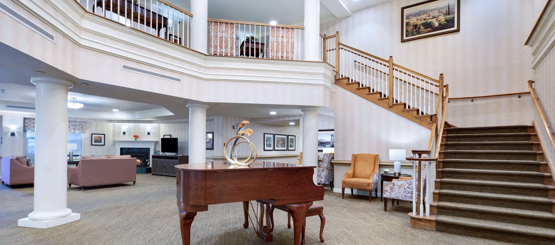 Lobby-style common room with a grand piano, seating areas, white columns, and a staircase leading to an upper balcony.