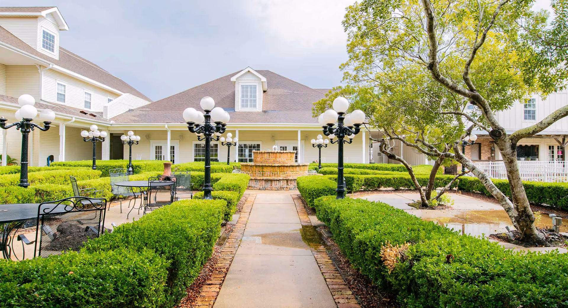 Outdoor courtyard area at Homestead Village Of Fairhope featuring a paved walkway lined with trimmed green hedges, black metal tables and chairs, multiple black lamp posts with white globes, a central tiered brick fountain, and surrounding buildings with beige siding and white trim under a cloudy sky.