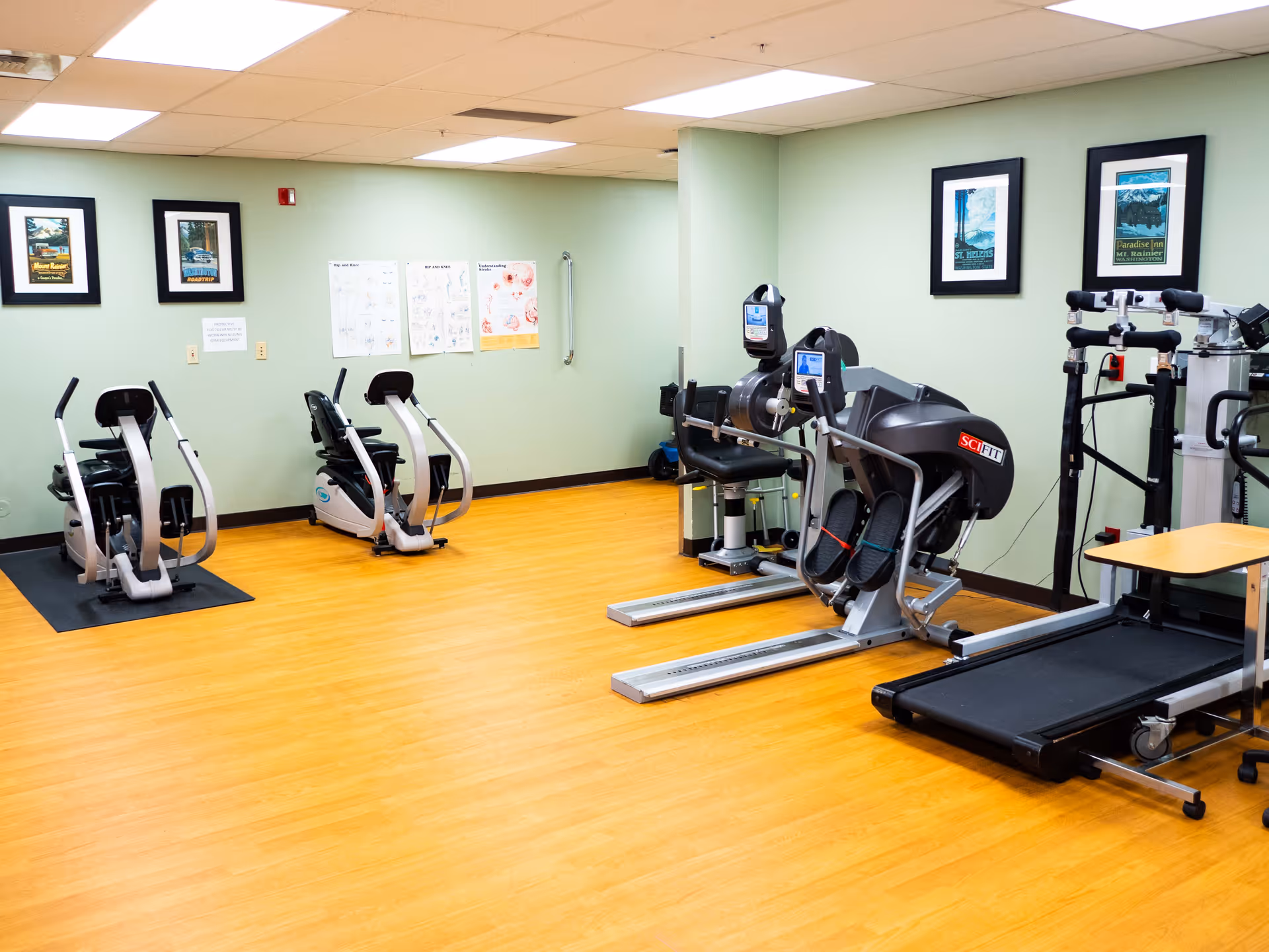A rehabilitation gym room with exercise equipment including two recumbent bikes, two SCI FIT machines, and a treadmill. The room has light green walls, wooden flooring, framed pictures on the walls, and instructional posters.